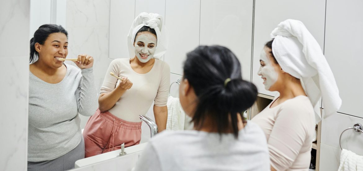 Two women smiling in a bathroom mirror while practicing daily dental care.